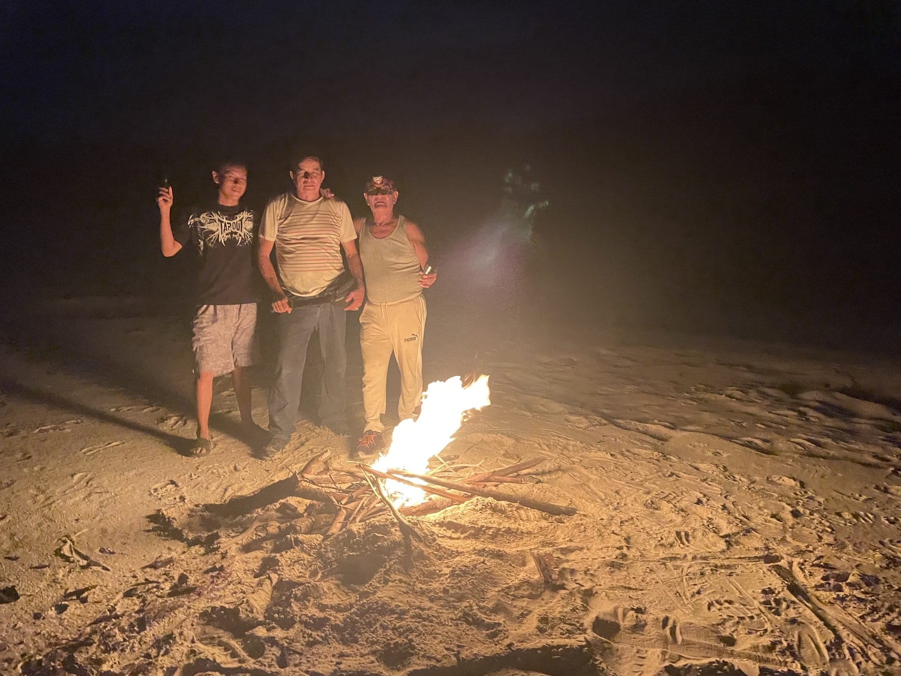 Guests gathered around a campfire on a white-sand river beach at night
